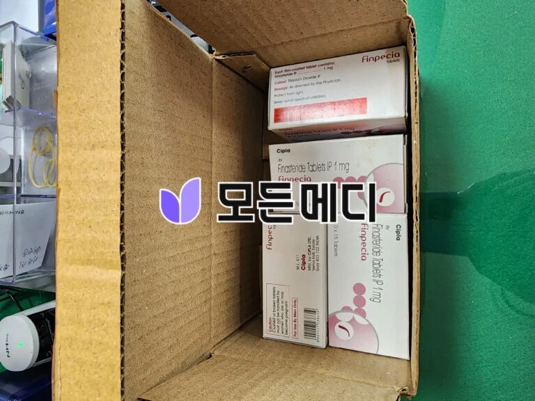 Open cardboard box containing pharmaceutical boxes labeled Finpecia finasteride tablets from Cipla, stacked inside against a green background.
