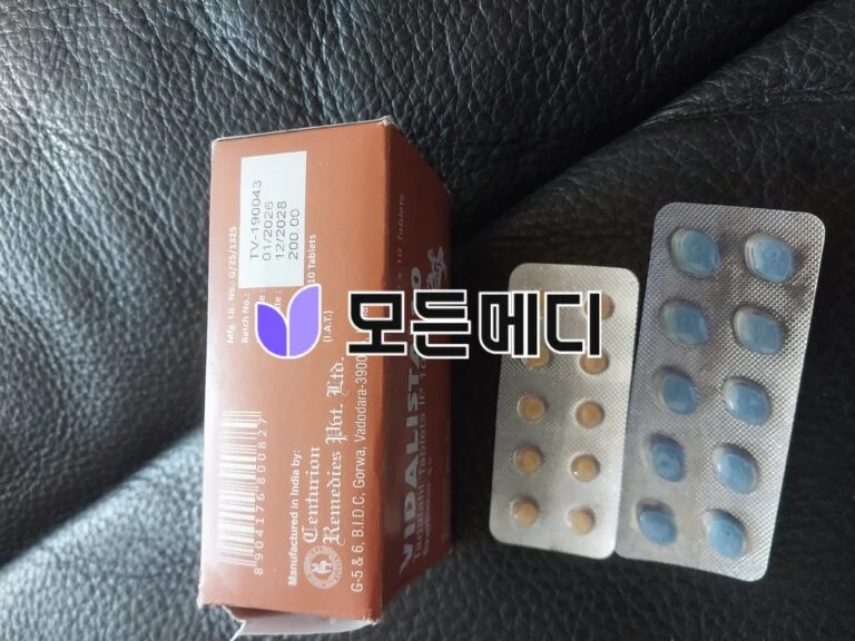 Close-up of a brown medicine box next to blister packs containing blue and yellow pills on a black leather surface, showing packaging details.