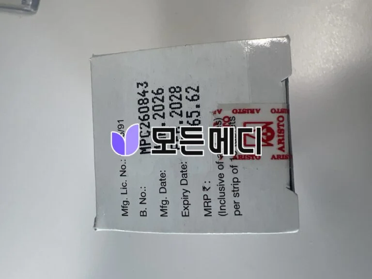 Close-up of a white cardboard box with black printed text and a red ARISTO label; a purple sticker partially covers the text.